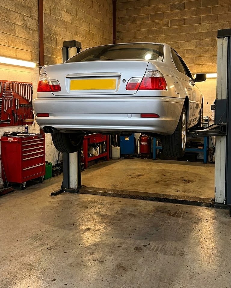 A silver car raised on a two post lift inside a tidy garage workshop, with tools on the wall and a red tool chest.