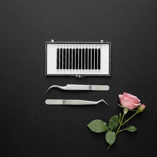 A tray of black lash extensions and two pairs of tweezers on a black surface, with one pink rose.