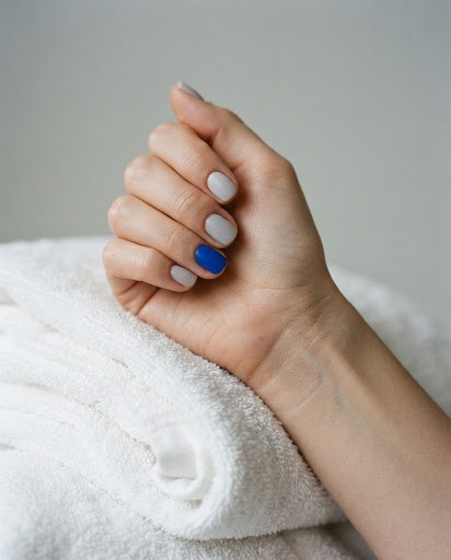 A hand resting on a folded white towel, with short pale grey nails and one cobalt blue nail.